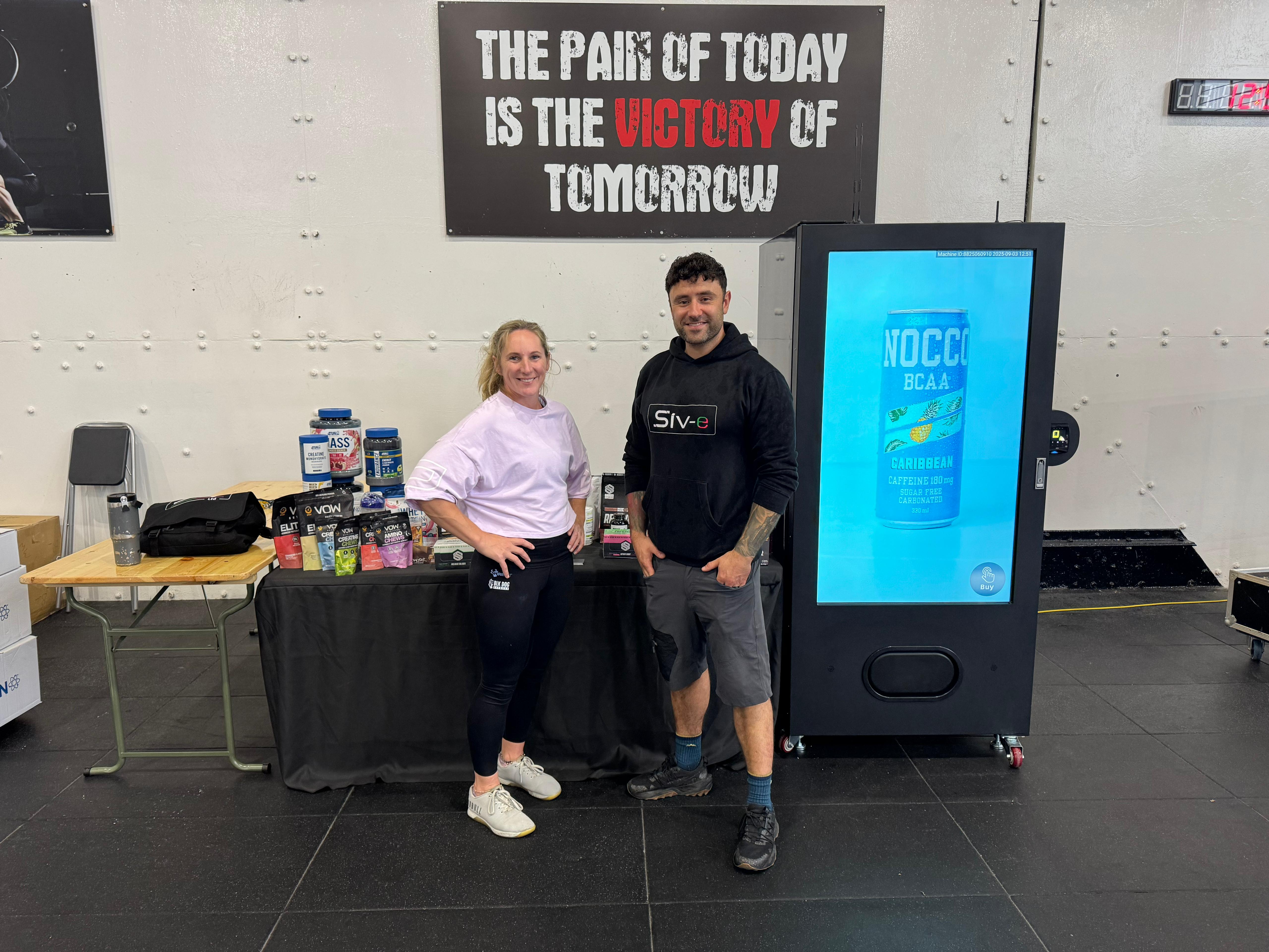 Two people beside an SIV vending machine and a sports nutrition product display in a gym.